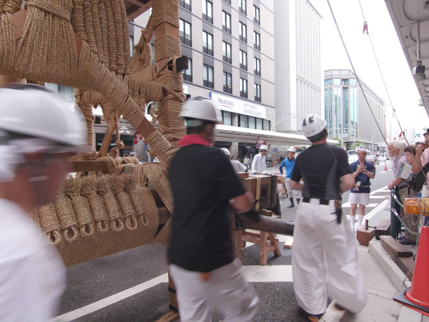 画像: 京都の祇園祭で大きな山鉾を準備する作業員たちが映っているシーン。町の通りに人々が集まり、伝統的な祭りの準備が