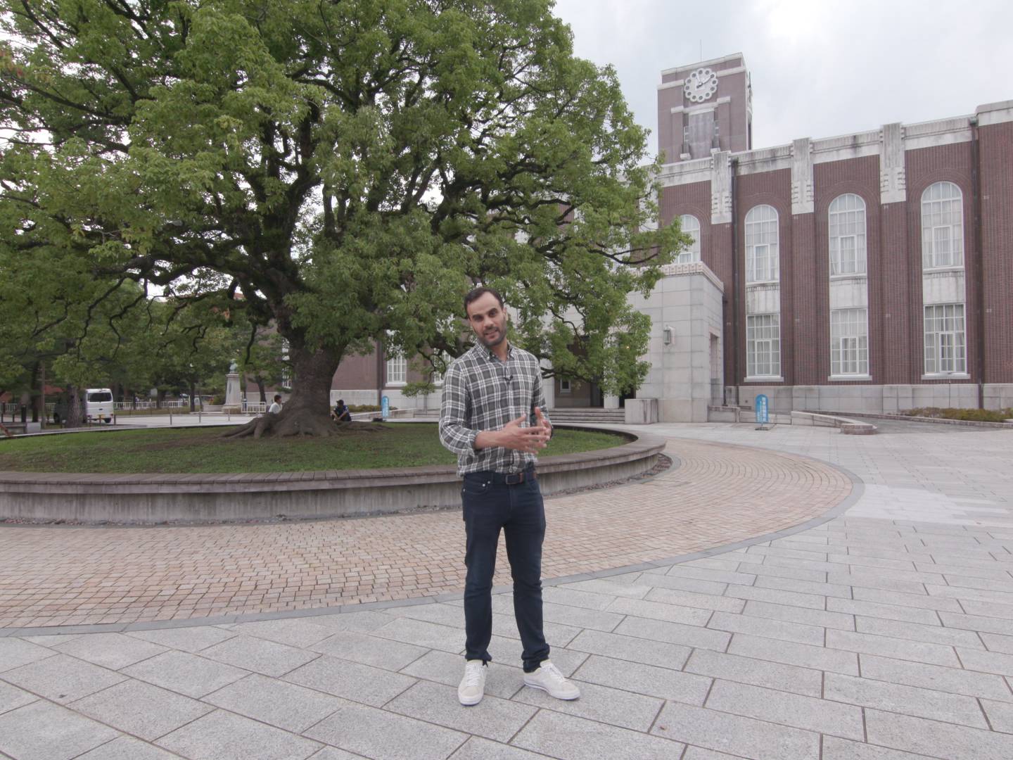 画像: 申し訳ありませんが、人物の特定や推測はできません。この画像は、大きな木と歴史的な建物が特徴的なキャンパスの風景を映しています。石畳の広場には