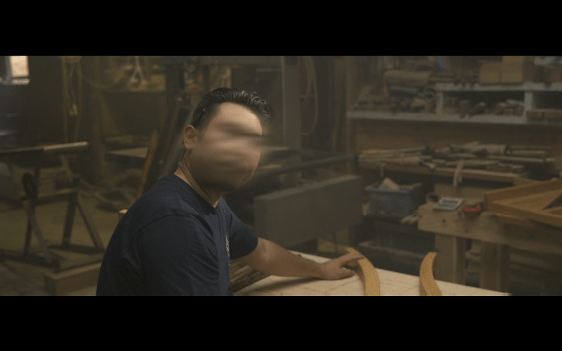 画像: 木工作業場で作業する人物が写っています。背景には様々な木工道具や材料が並んでおり、実用的な木工環境が正確に描かれています。工房
