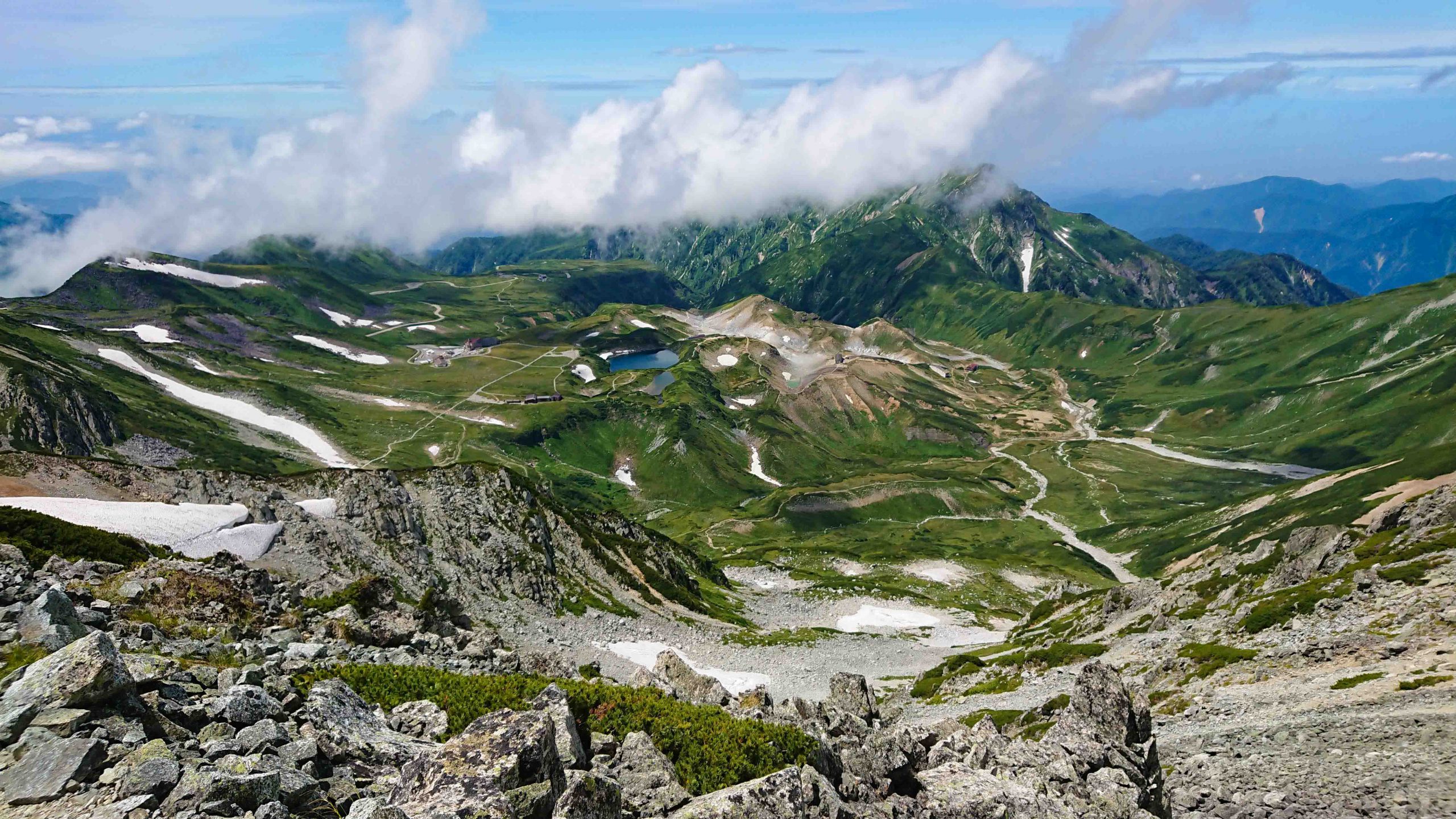 画像: この画像は、立山連峰の美しい風景を捉えています。青空の下に広がる緑豊かな山々と谷、そして雪が残る山肌が特徴的です。登山