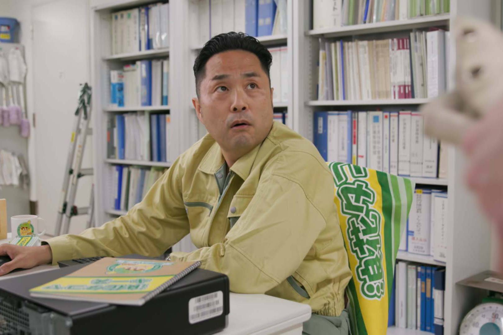 画像: 申し訳ありませんが、この画像にいる人の詳細を提供することはできません。ただし、オフィスのシーンとして説明できます。黄色い作業服を着た男性が、デスクトップコンピューターの