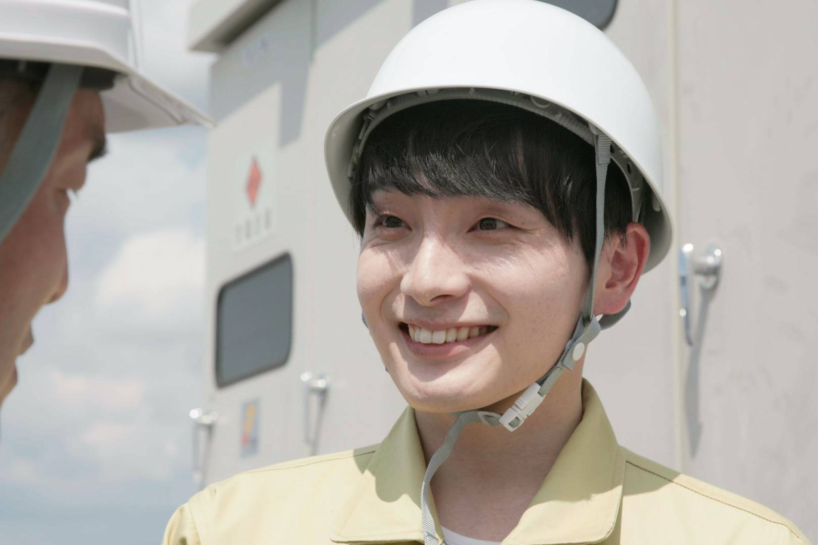 画像: 申し訳ありませんが、この画像の人物について特定することはできません。安全帽を着用した作業員が笑顔で、屋外の工事現場にいる様子が描かれています。背景には