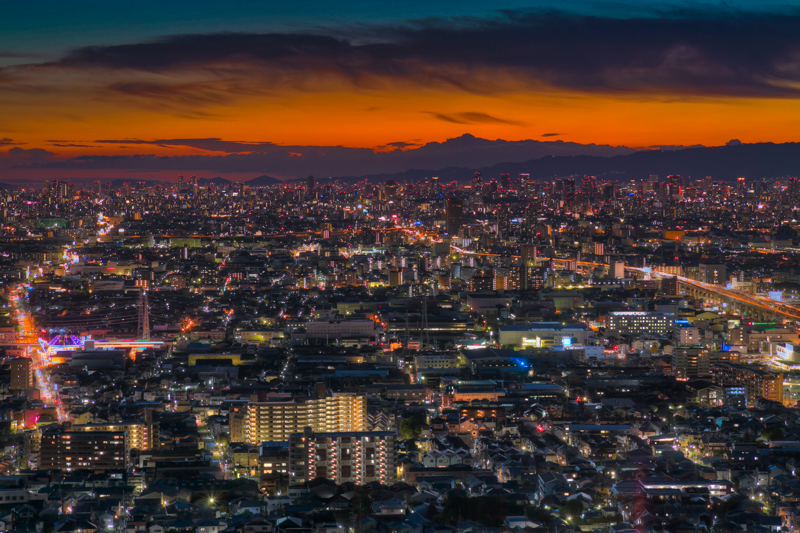 画像: 夜景の大阪市街地のパノラマビュー。夕焼けが鮮やかに空を彩り、ビルや街灯が街を輝かせています。都会の活気と静けさ