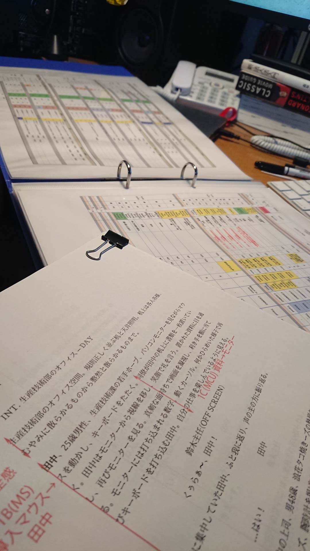 画像: オフィスのデスクに置かれたクリップ付きの書類や図がまとめられているバインダーが見えます。書類にはスケジュールや計画表が表示されています。キーワード: