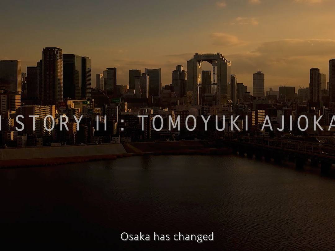 画像: 大阪の夕暮れに映える高層ビル群を背景に、「KIKUICHI STORY II : TOMOYUKI AJIOKA, GRIND」のテキストが表示されている画像。都市の変化