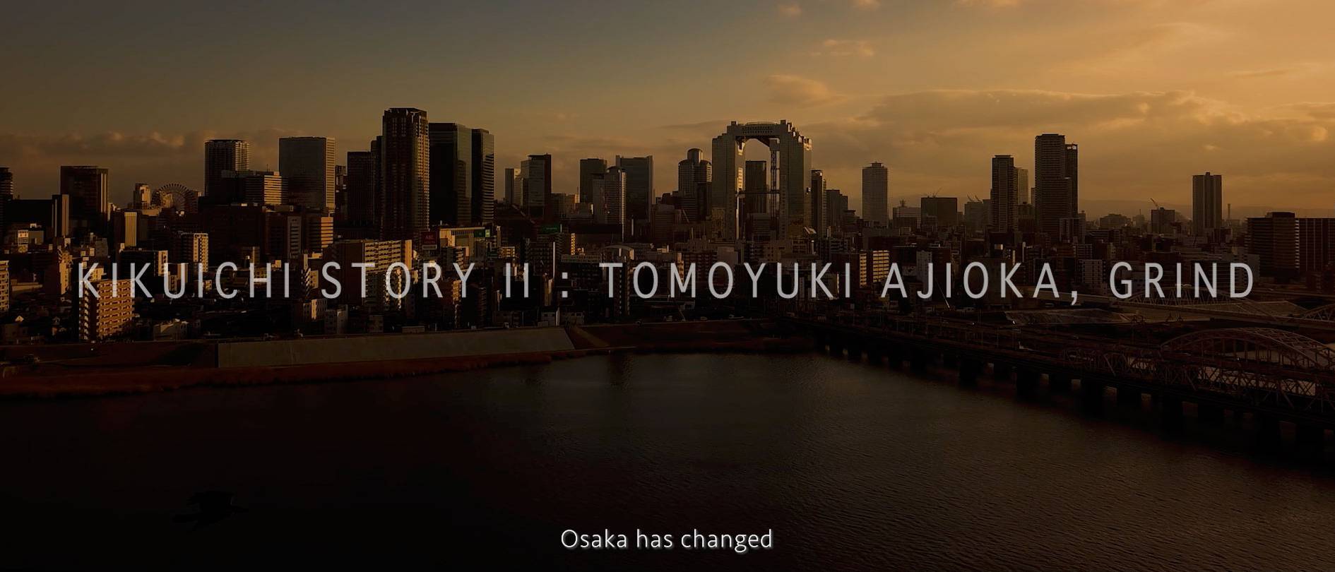 画像: 大阪の夕暮れに映える高層ビル群を背景に、「KIKUICHI STORY II : TOMOYUKI AJIOKA, GRIND」のテキストが表示されている画像。都市の変化