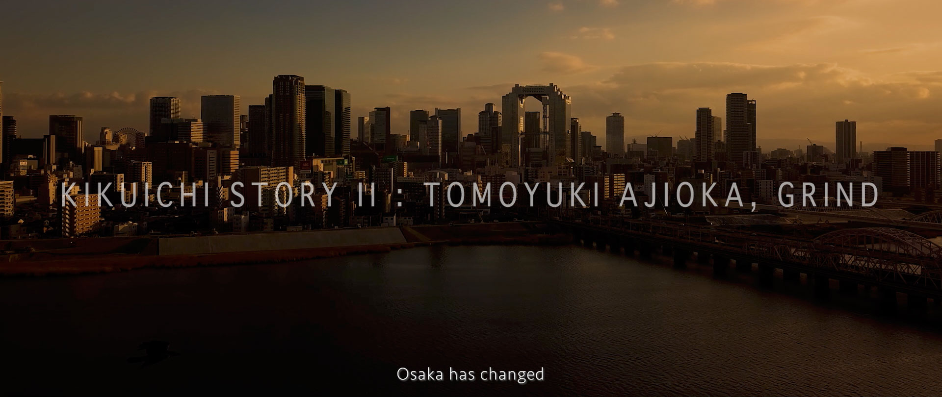 画像: 大阪の夕暮れに映える高層ビル群を背景に、「KIKUICHI STORY II : TOMOYUKI AJIOKA, GRIND」のテキストが表示されている画像。都市の変化