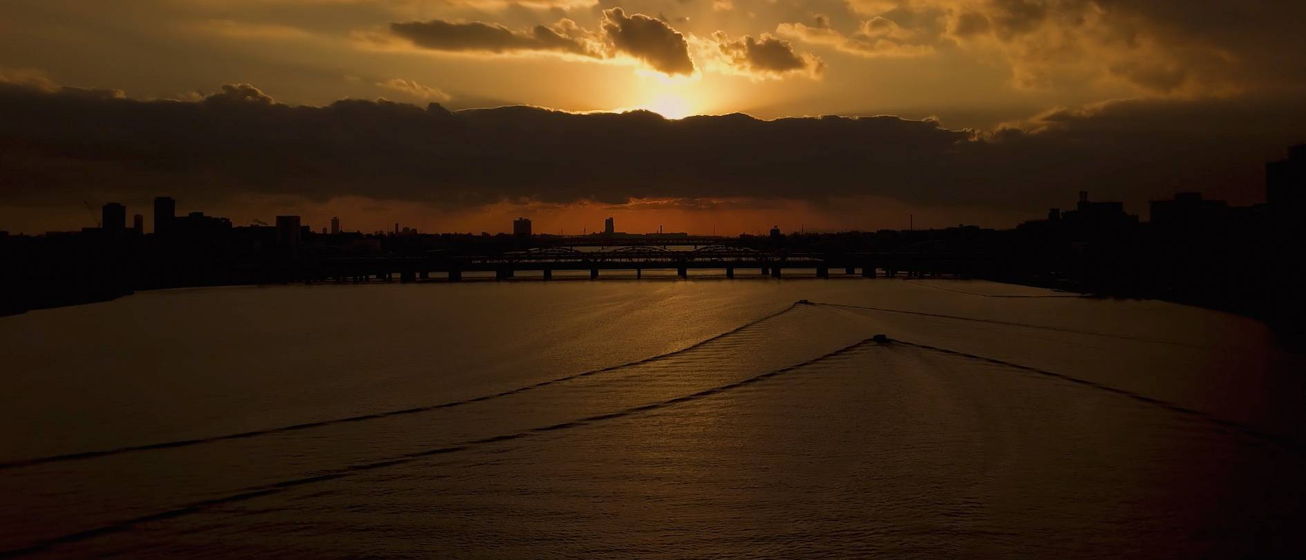 画像: 夕日が川の上に沈む光景。シルエットの橋とビル群が薄暗い空を背景に現れ、水面にはボートの波紋が広がっています。キーワード: